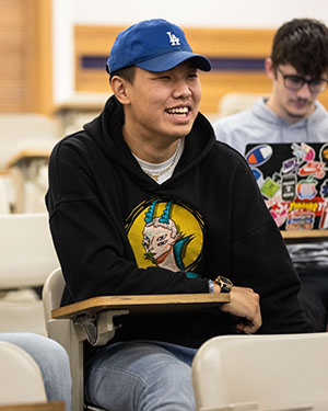 A student sits at a desk. They are wearing a hoodie and baseball cap and smiling while looking off to the right