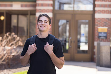A student stands outside in a black t-shirt and backpack. They are holding the handles of the backpack and smiling.
