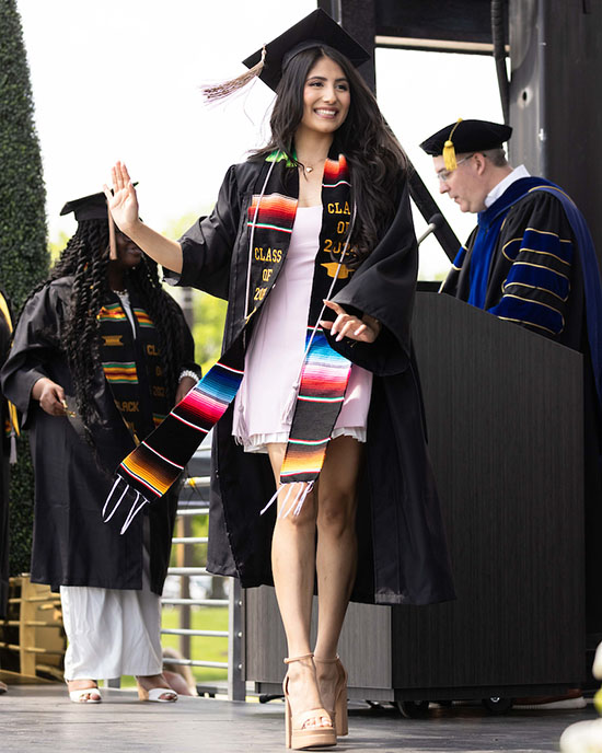  A student walks across stage to get their diploma. They are wearing a Latino affinity stole