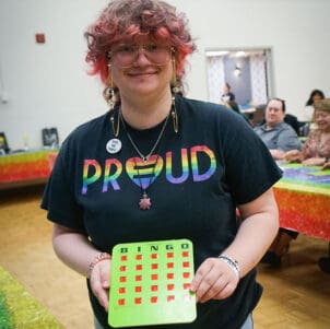 A student holds up a completed bingo card and smiles. They are wearing a rainbow shirt that reads "Proud"
