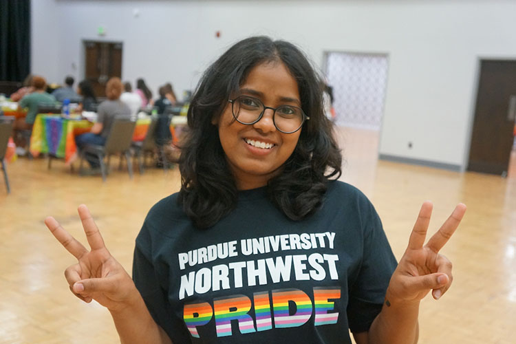 A student holds up double peace signs and smiles. They are wearing a black PNW Pride shirt