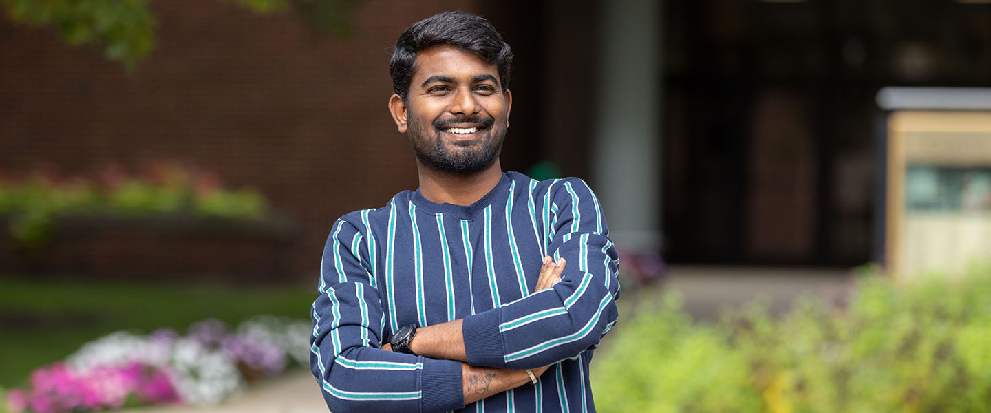 A student in a blue striped long sleeve shirt crosses their arms and smiles