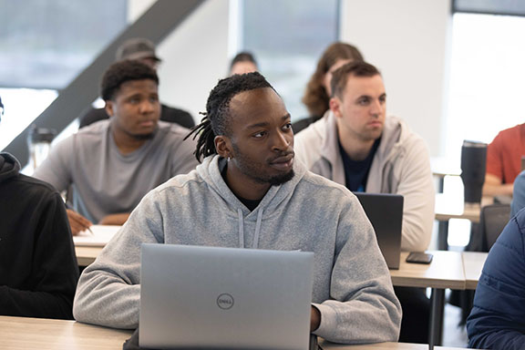 A student sits at a table in a classroom. There is a laptop open on the table in front of them