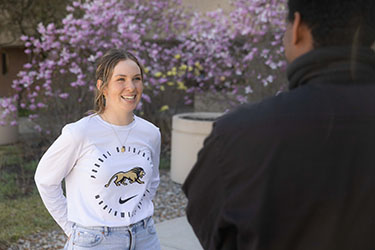 A student in a white long sleeve athletics t-shirt smiles outside on campus.