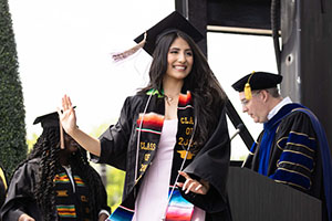 A student in commencement regalia and Hispanic affinity stole walks across a stage