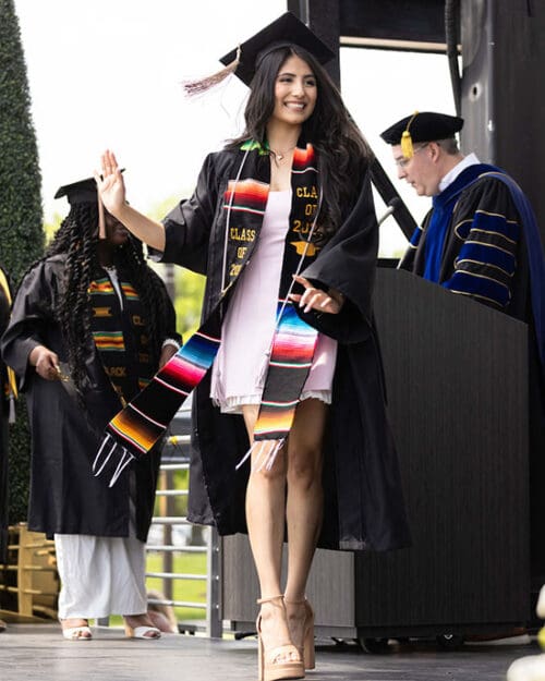 A student in commencement regalia and Hispanic affinity stole walks across a stage