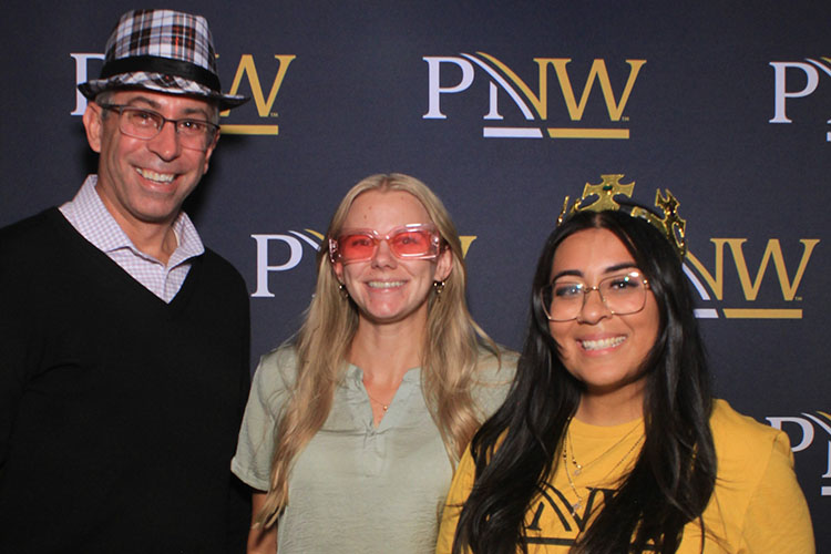 Three people pose together in front of a PNW branded backdrop. The people in the photo are wearing the following props (left to right): a plaid hat, red sunglasses and a gold crown