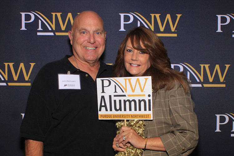 Two people pose in front of a PNW branded backdrop. The person on the right is holding up a "PNW Alumni" sign