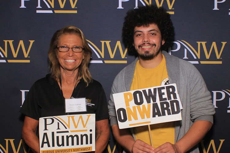 Two people stand side by side in front of a PNW branded backdrop. The person on the left is holding a "PNW Alumni" sign and the person on the right has a "Power Onward" sign