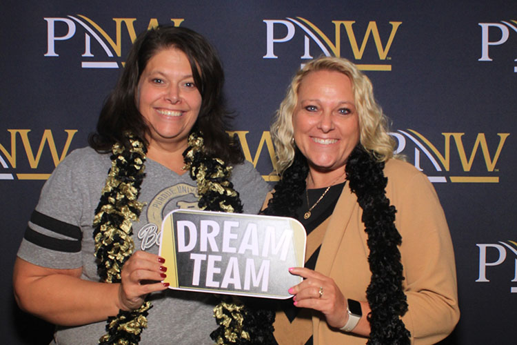 Two people hold up a sign that says "Dream Team". They are both wearing feather boas and standing in front of a PNW branded backdrop