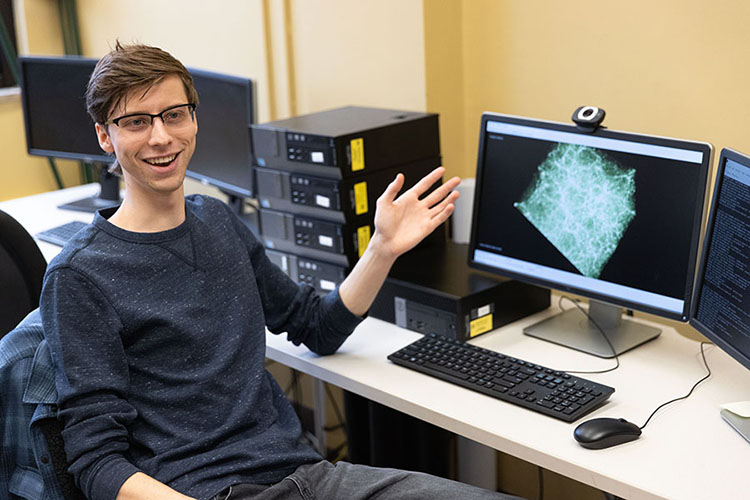 A student sits at a computer desk. He is looking at the camera and gesturing toward a graphic on the screen.