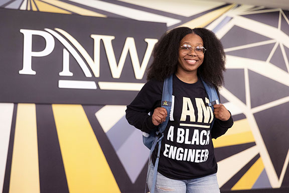 A female student wearing an "I am a black engineer" shirt stands in front of a wall with a PNW logo.