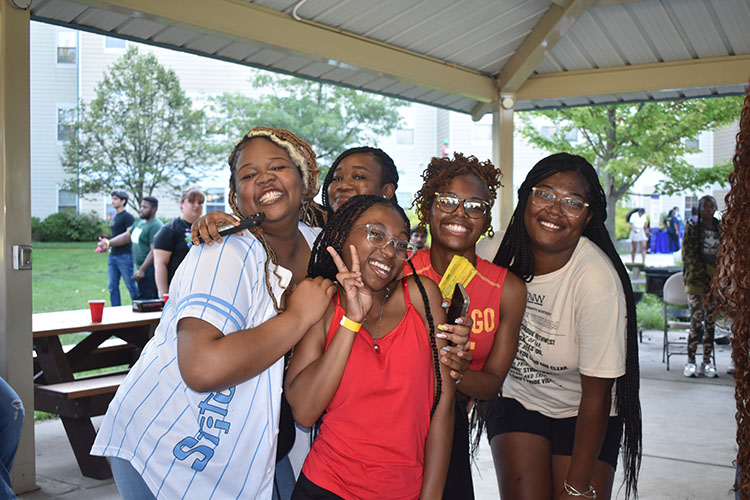 Five students lean into each other and smile at a camera. There are more people in the background of the photo