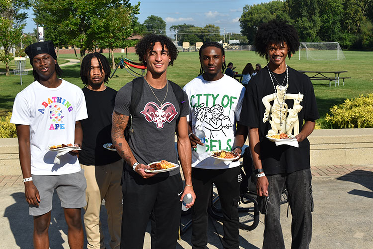 Five students pose together outside. They are holding plates of food