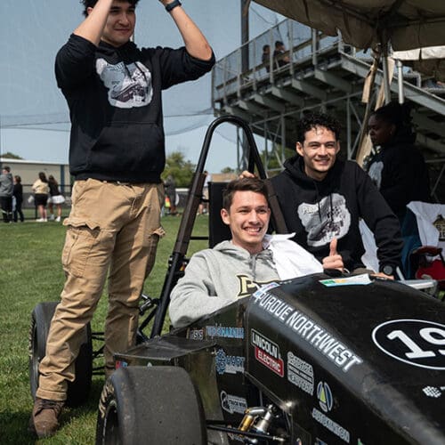 Students around a baja car. One student is sitting in the drivers seat, another is crouched down and leaning against the car, and the third is standing behind the car.