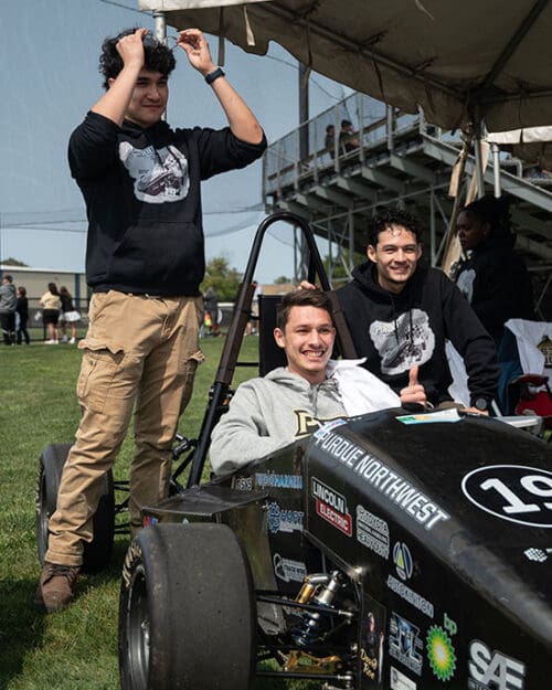 Students around a baja car. One student is sitting in the drivers seat, another is crouched down and leaning against the car, and the third is standing behind the car.