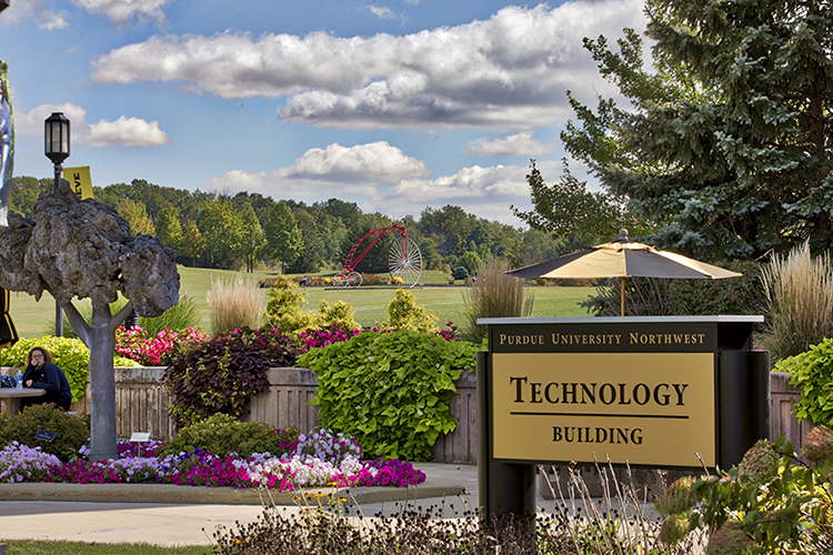 A sign on the Westville Campus reading "Purdue University Northwest Technology Building". The campus greenery and an umbrella are in the background.