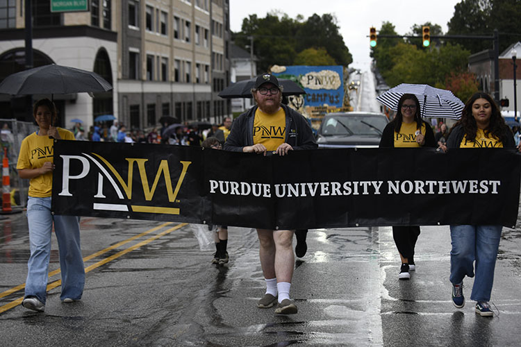 Several people walk behind a long black banner that has the PNW logo and reads "Purdue University Northwest"