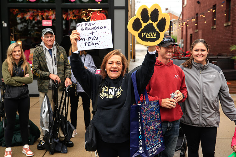 A person a black PNW sweatshirt holds up a sign in their right hand that reads "My Fav Grandson + My Fav Team" and on their left hand, they have a foam PNW "paw"