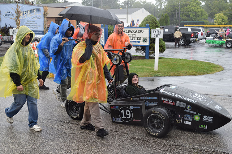 Several students wear rain ponchos and walk near a racecar. One of them is holding an umbrella