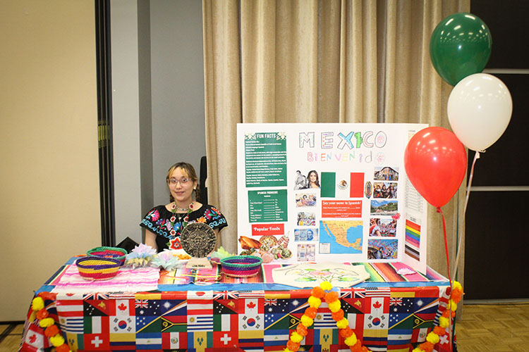 a student in traditional Mexican clothing sits at a "Mexico" table