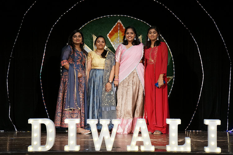 Four students in traditional Indian saris stand behind a marquee Diwali sign