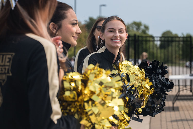 PNW cheerleaders smile and hold black and gold pompoms. One of the cheerleaders is leaning out of line and smiling at the camera