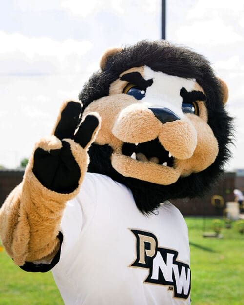 PNW lion mascot Leo raises two fingers at PNW's outdoor Dowling Park field.