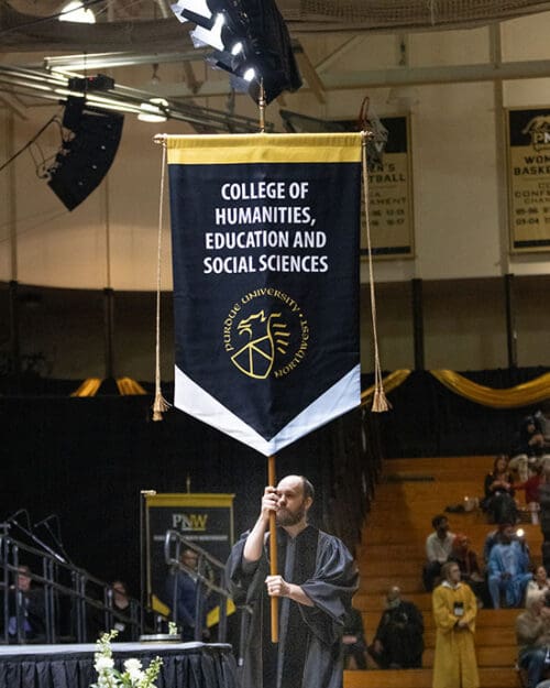 A graduate marches with a College of Humanities, Education and Social Sciences banner at PNW commencement