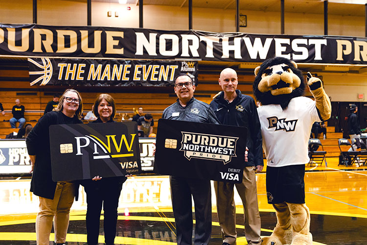 Several people hold up mock Purdue University Northwest credit cards beside PNW mascot Leo center court at a basketball game.